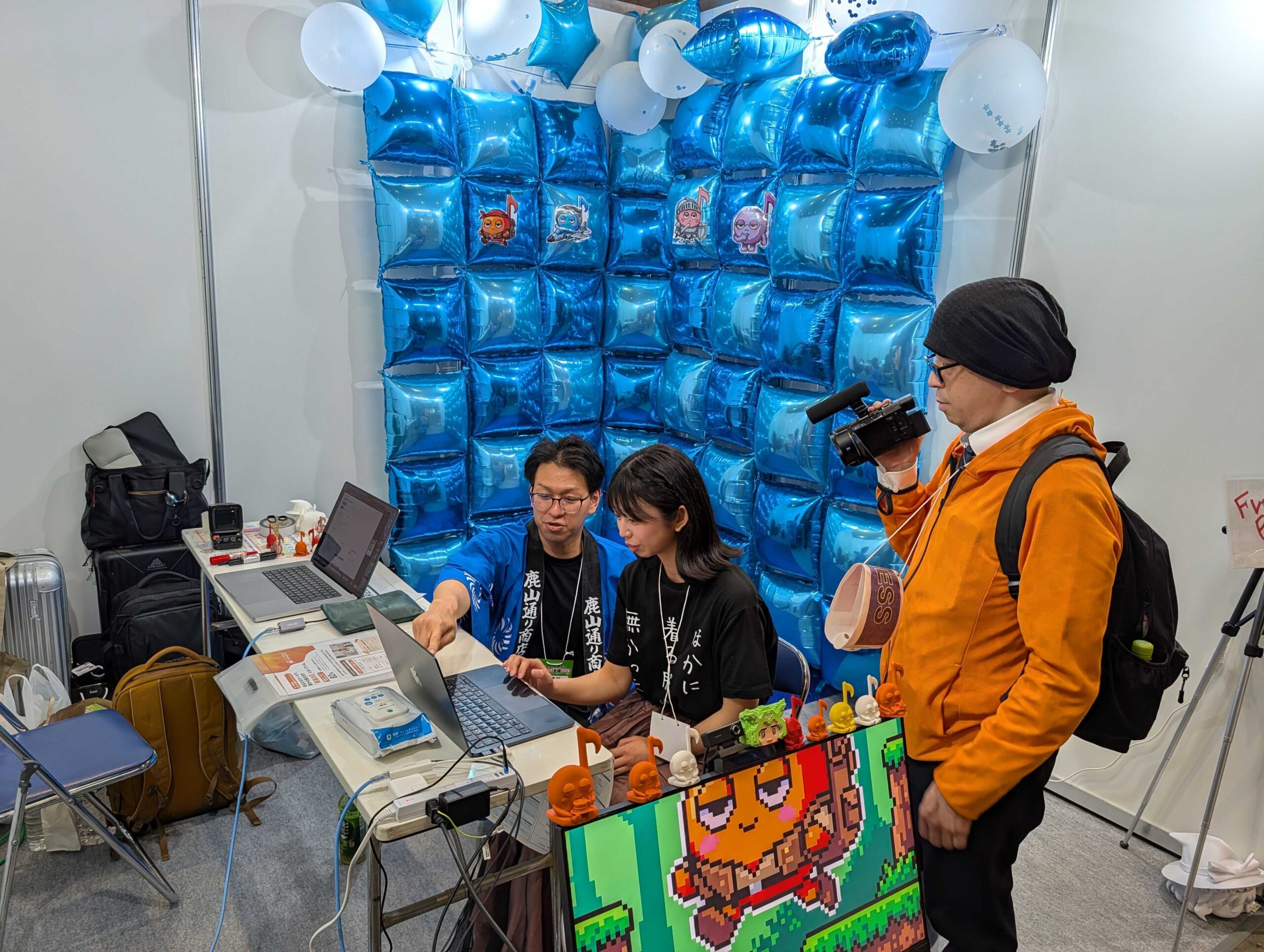 ニコニコ超会議2026 がだんだん株式会社ブース全景。青いバルーンの背景、ゲームキャラクターのフィギュア、メディアによる取材の様子